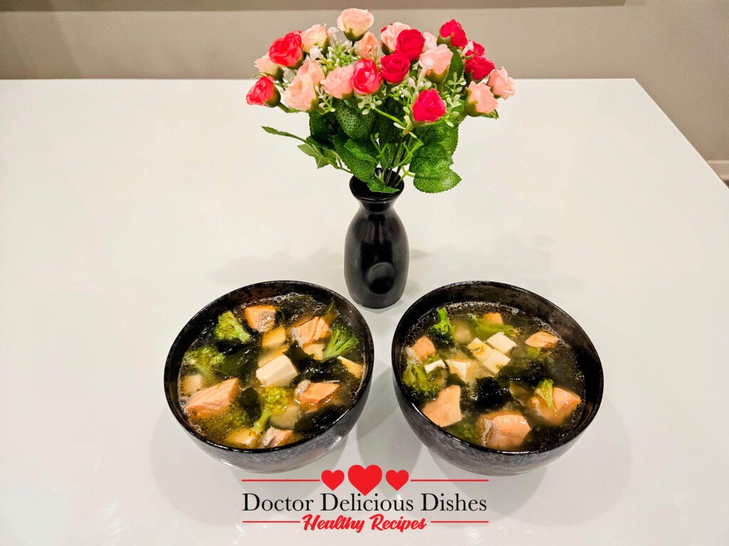 Two bowls of Salmon Tofu Miso Soup on a clean table setting, adorned with a vase of colorful flowers.