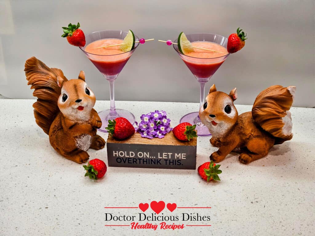 Two Strawberry, Banana, Orange Juice, smoothies in purple martini glasses with cute decorations of squirrel statues and fruit with a small sign that says "HOLD ON... LET ME OVERTHINK THIS".