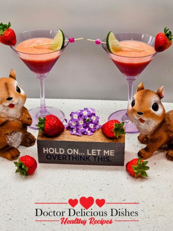 Two Strawberry, Banana, Orange Juice, smoothies in purple martini glasses with cute decorations of squirrel statues and fruit with a small sign that says "HOLD ON... LET ME OVERTHINK THIS".