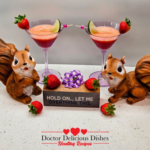 Two Strawberry, Banana, Orange Juice, smoothies in purple martini glasses with cute decorations of squirrel statues and fruit with a small sign that says "HOLD ON... LET ME OVERTHINK THIS".