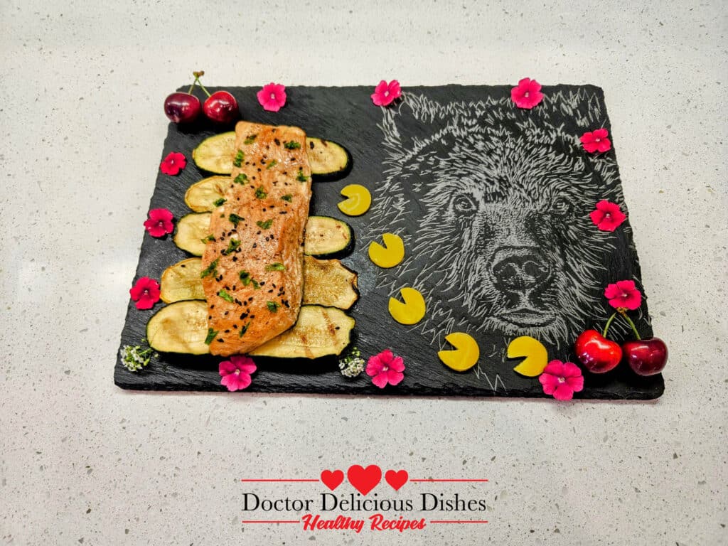 Close-up of the plated grilled Kewpie salmon on a slate board with grilled zucchini slices, garnished with black sesame seeds, cherries, and edible flowers, showcasing the vibrant presentation.