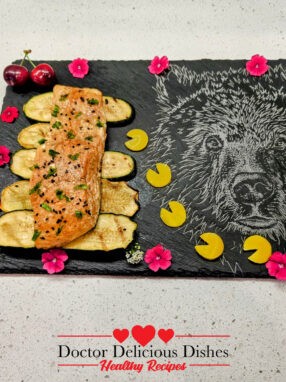 Close-up of the plated grilled Kewpie salmon on a slate board with grilled zucchini slices, garnished with black sesame seeds, cherries, and edible flowers, showcasing the vibrant presentation.