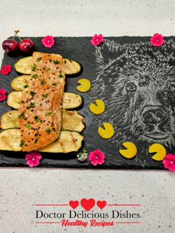 Close-up of the plated grilled Kewpie salmon on a slate board with grilled zucchini slices, garnished with black sesame seeds, cherries, and edible flowers, showcasing the vibrant presentation.