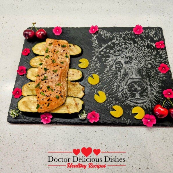 Close-up of the plated grilled Kewpie salmon on a slate board with grilled zucchini slices, garnished with black sesame seeds, cherries, and edible flowers, showcasing the vibrant presentation.