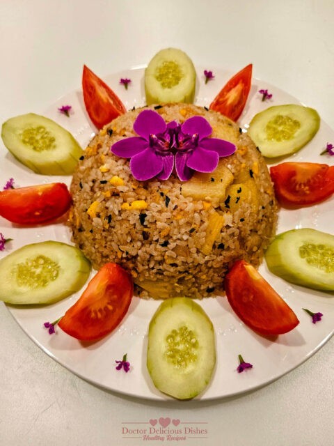 Colorful and beautifully arranged Thai fried rice with cod, garnished with slices of cucumber, wedges of tomato, and vibrant purple edible flowers on a white circular plate.