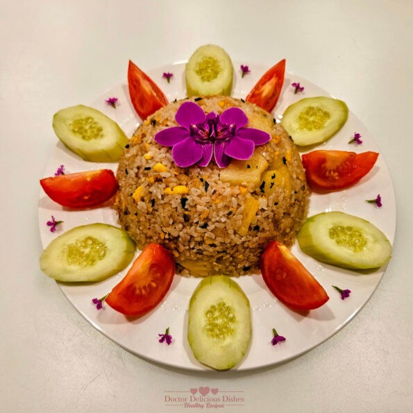 Colorful and beautifully arranged Thai fried rice with cod, garnished with slices of cucumber, wedges of tomato, and vibrant purple edible flowers on a white circular plate.