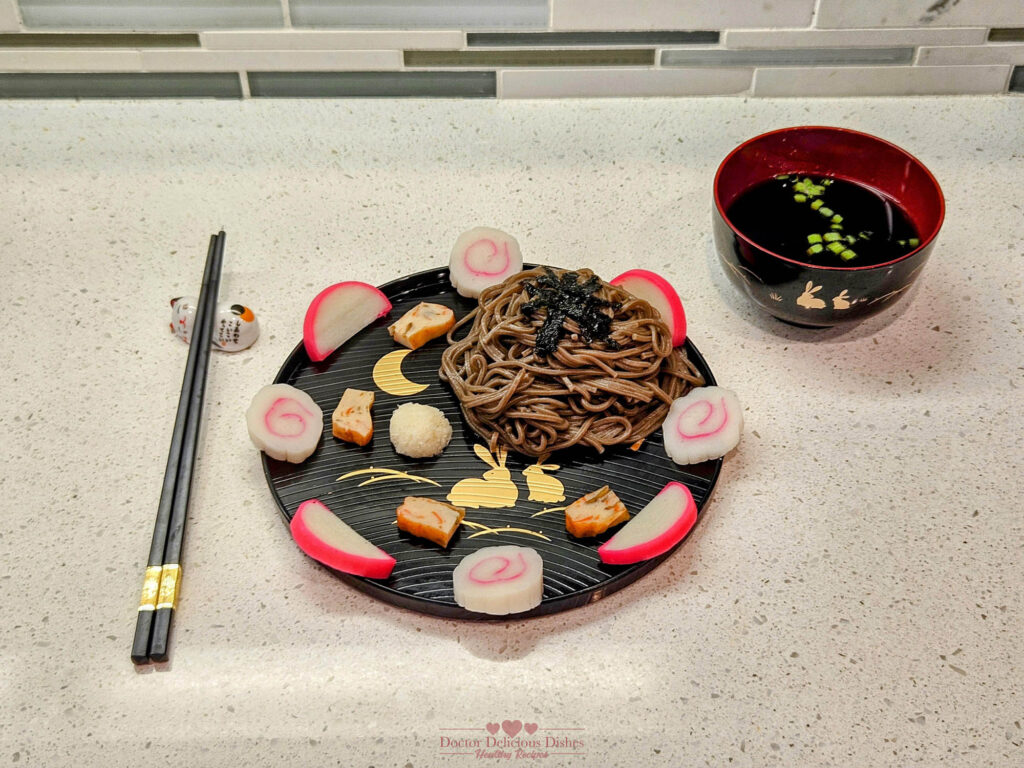 A beautifully arranged plate of cold soba noodles with fish cake, garnished with seaweed, daikon radish, and green onions. The dish is artfully presented on a black plate with golden patterns, accompanied by a bowl of soup and a pair of black chopsticks, showcasing a balanced and nutritious Japanese meal.