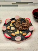 A beautifully arranged plate of cold soba noodles with fish cake, garnished with seaweed, daikon radish, and green onions. The dish is artfully presented on a black plate with golden patterns, accompanied by a bowl of soup and a pair of black chopsticks, showcasing a balanced and nutritious Japanese meal.