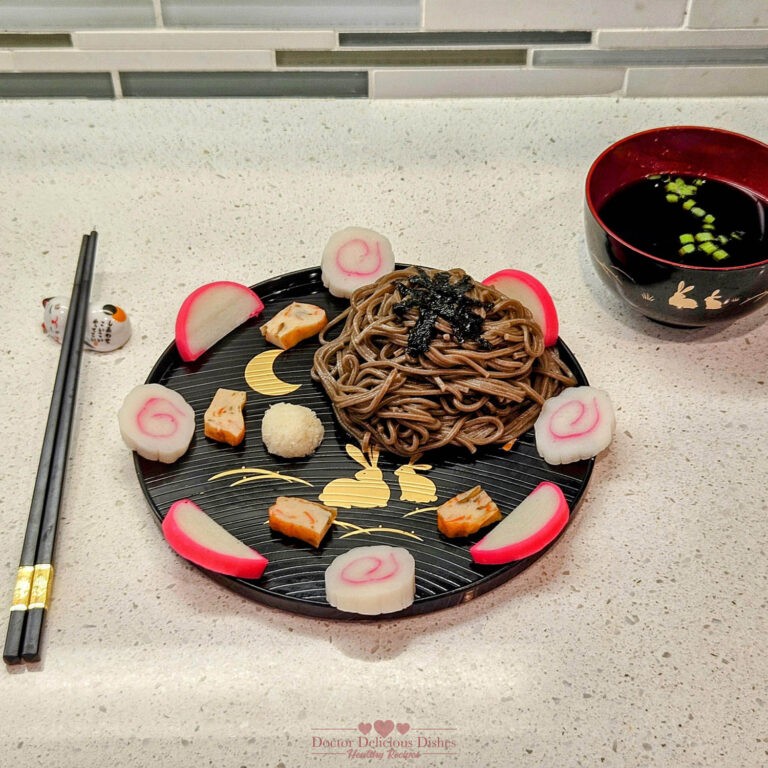 A beautifully arranged plate of cold soba noodles with fish cake, garnished with seaweed, daikon radish, and green onions. The dish is artfully presented on a black plate with golden patterns, accompanied by a bowl of soup and a pair of black chopsticks, showcasing a balanced and nutritious Japanese meal.