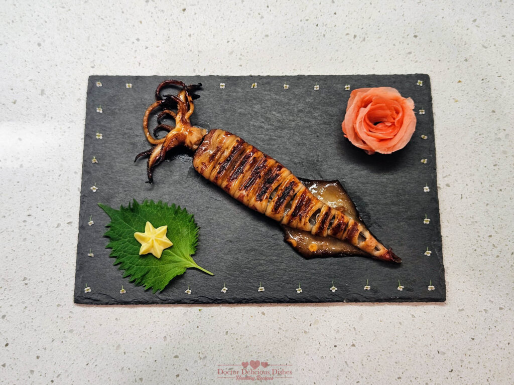 Grilled squid skewer served on a decorative slate plate with shiso leaf, star-shaped tamagoyaki, and a rose garnish, creating an elegant presentation.