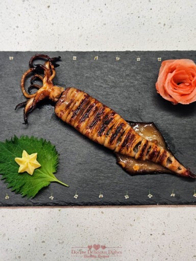 Grilled squid skewer served on a decorative slate plate with shiso leaf, star-shaped tamagoyaki, and a rose garnish, creating an elegant presentation.