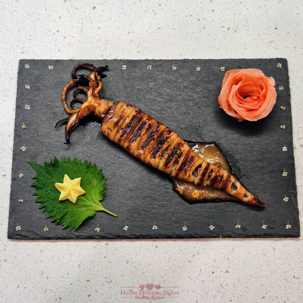 Grilled squid skewer served on a decorative slate plate with shiso leaf, star-shaped tamagoyaki, and a rose garnish, creating an elegant presentation.