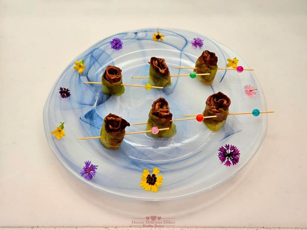 Cucumber Wrap with Beef Short Rib Bites presented on a decorative white and blue plate with colorful flower garnishes. Each bite is secured with a wooden skewer adorned with colorful beads.