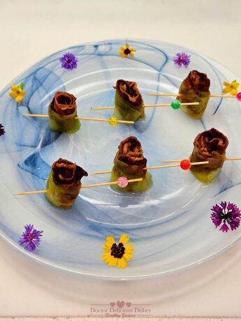 Cucumber-Wrapped Beef Short Rib Bites presented on a decorative white and blue plate with colorful flower garnishes. Each bite is secured with a wooden skewer adorned with colorful beads.