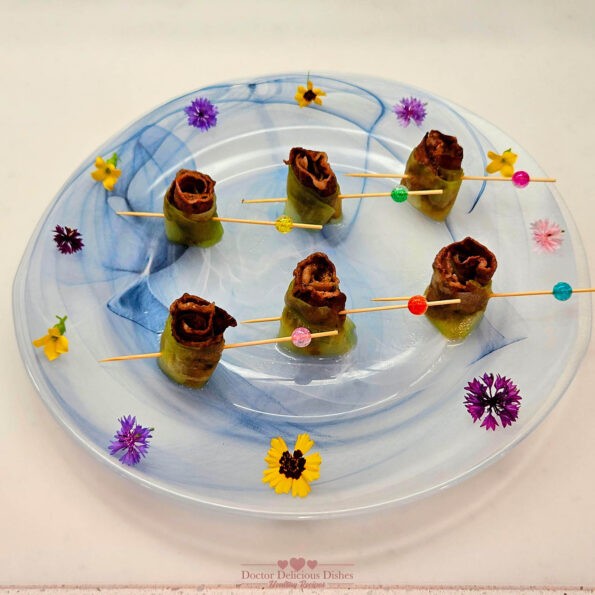 Cucumber-Wrapped Beef Short Rib Bites presented on a decorative white and blue plate with colorful flower garnishes. Each bite is secured with a wooden skewer adorned with colorful beads.