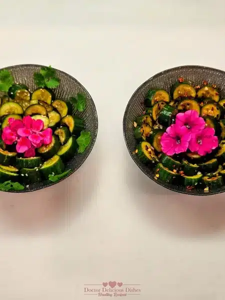 Two bowls of Chinese Cucumber Salad garnished with pink flowers and cilantro, presented side by side, with one spicy and one non-spicy version, showcasing vibrant and fresh ingredients.