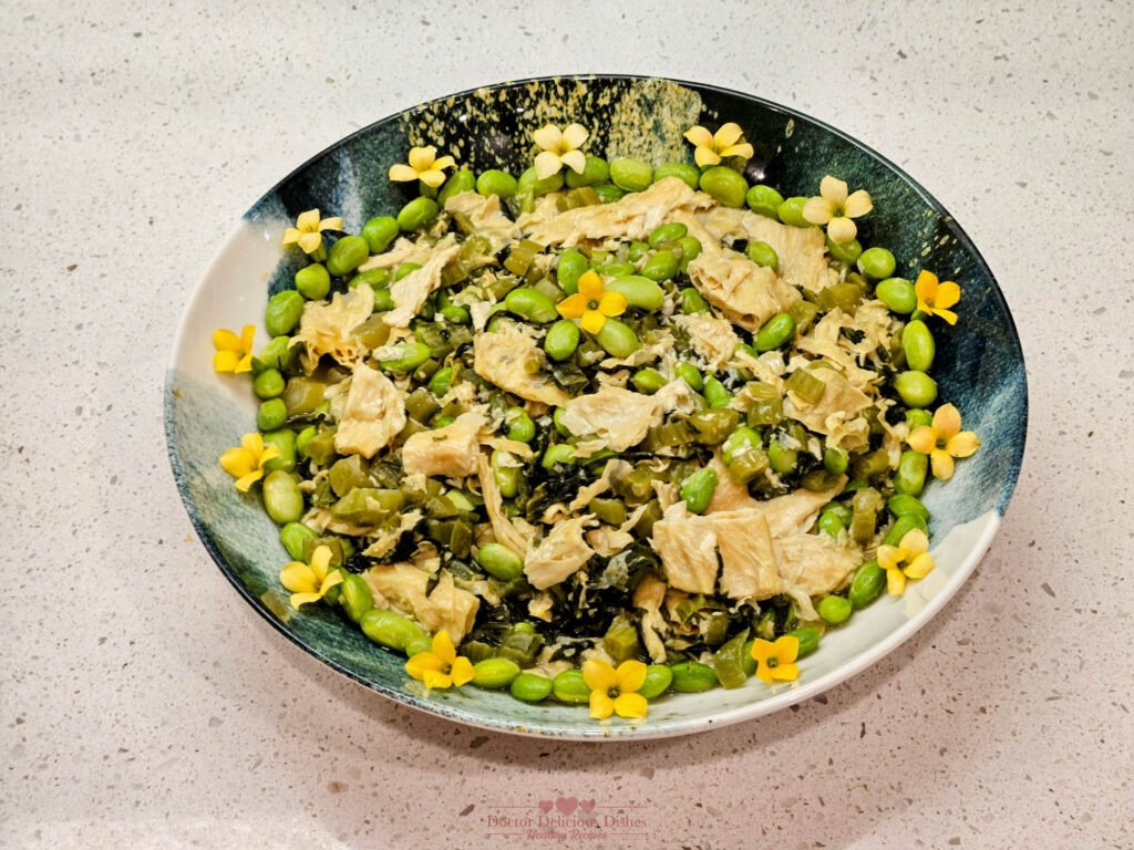 A vibrant Chinese dish featuring tofu skin, edamame, and preserved mustard greens, garnished with yellow blossoms, beautifully arranged in a blue and white asian style bowl.