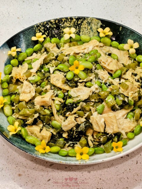 A vibrant Chinese dish featuring tofu skin, edamame, and preserved mustard greens, garnished with yellow blossoms, beautifully arranged in a blue and white asian style bowl.