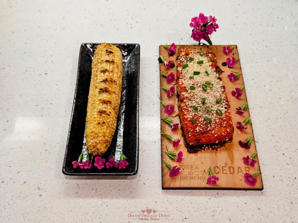 Final presentation of the grilled cedar plank teriyaki salmon garnished with green onions, served alongside grilled Japanese-style corn on the cob, both beautifully arranged with edible flowers on a serving board and plate.