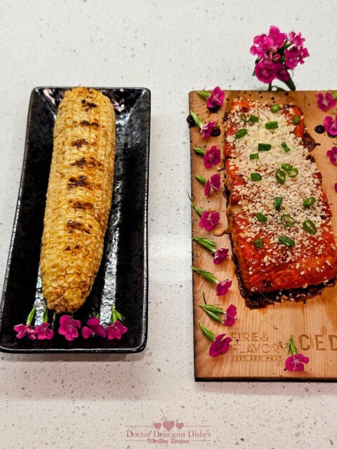 Final presentation of the grilled cedar plank teriyaki salmon garnished with green onions, served alongside grilled Japanese-style corn on the cob, both beautifully arranged with edible flowers on a serving board and plate.