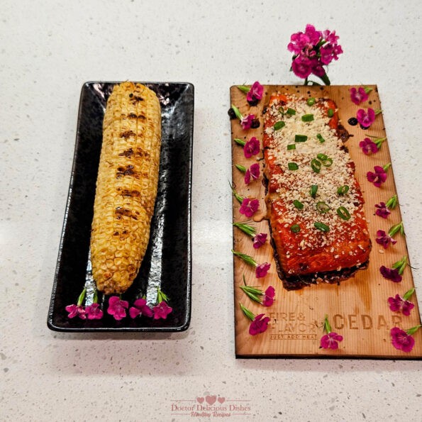 Final presentation of the grilled cedar plank teriyaki salmon garnished with green onions, served alongside grilled Japanese-style corn on the cob, both beautifully arranged with edible flowers on a serving board and plate.