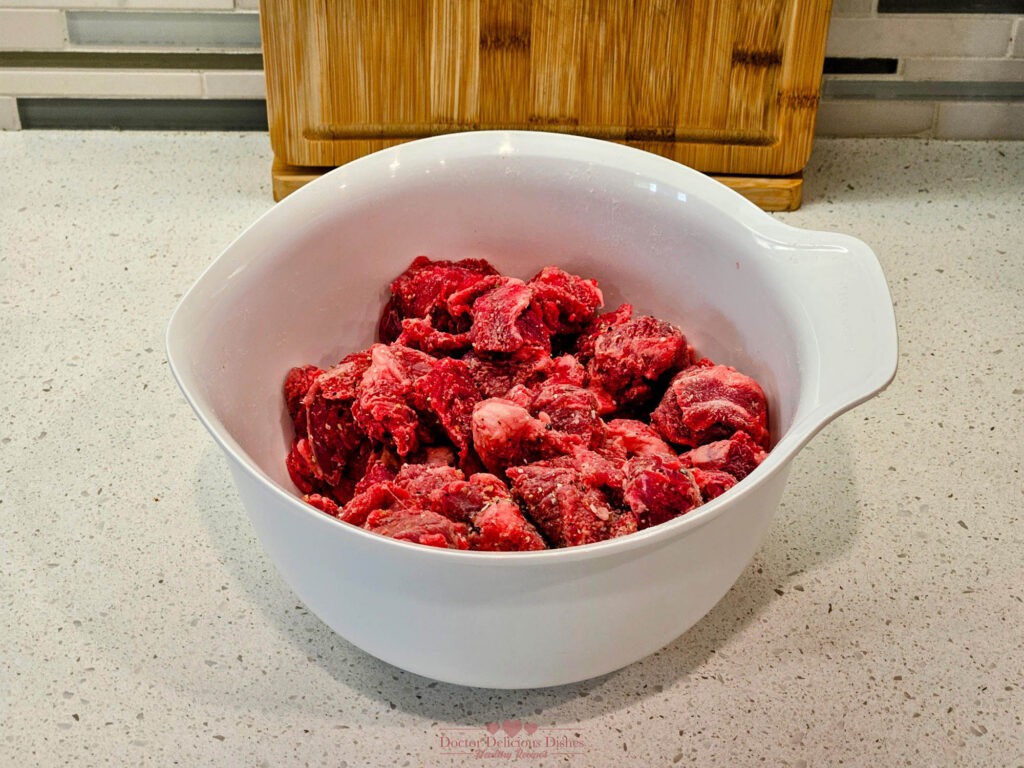 Beef cubes seasoned with salt, pepper, and garlic powder in a white mixing bowl, ready for browning.