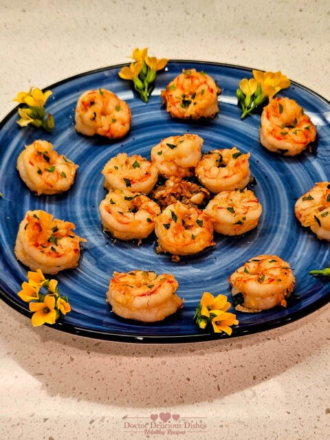 A beautifully arranged platter of Garlic Shrimp, garnished with parsley, oregano, basil and decorative yellow flowers on a blue plate.