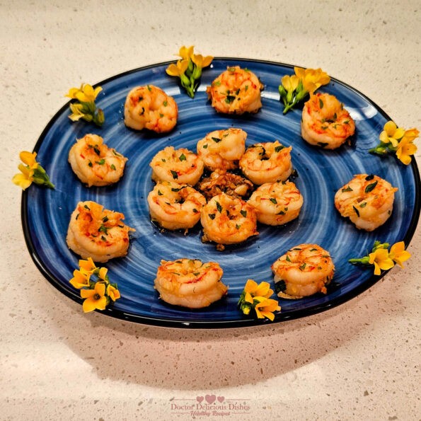 A beautifully arranged platter of Garlic Shrimp, garnished with parsley, oregano, basil and decorative yellow flowers on a blue plate.