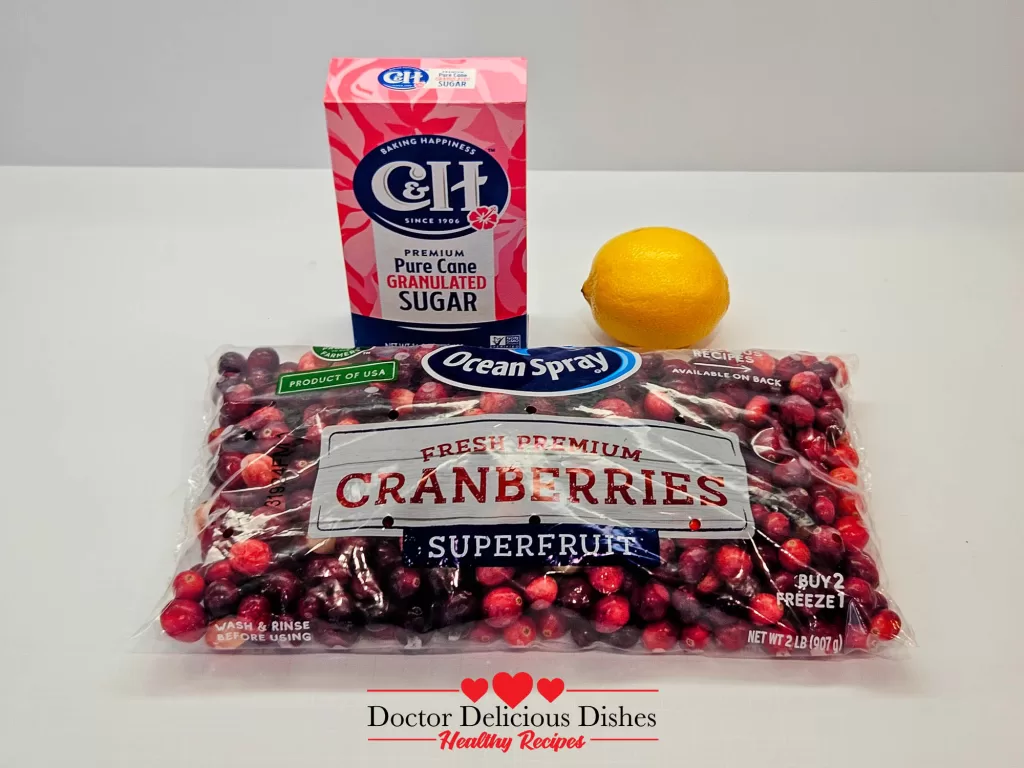 Fresh cranberries, a package of sugar, and a bright yellow lemon arranged on a white surface, illustrating the simple ingredients.