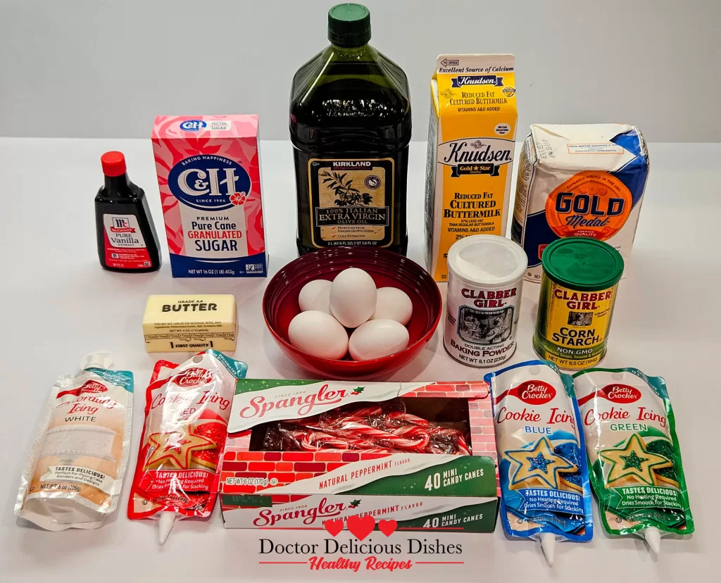 An assortment of baking ingredients neatly arranged on a white surface, featuring items like sugar, vanilla, olive oil, butter, eggs, cornstarch, and buttermilk along with packaged candy canes.