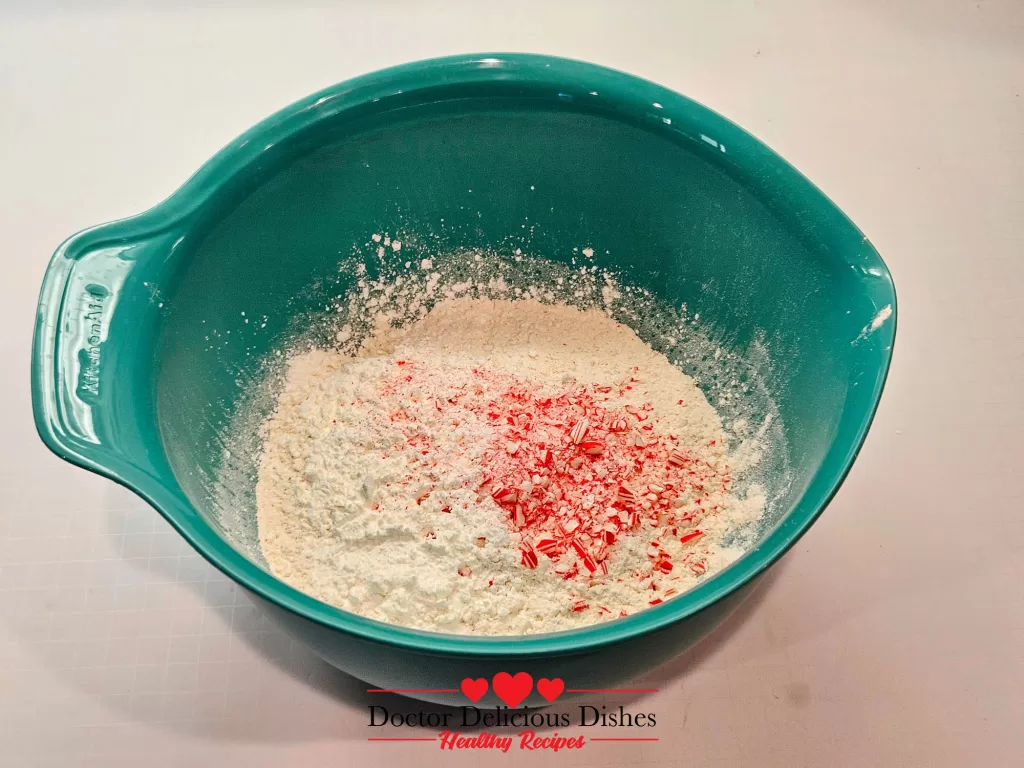 A turquoise mixing bowl containing a flour mixture with vibrant red crushed peppermint candy canes sprinkled on top, ready to be gently folded into the dry ingredients.