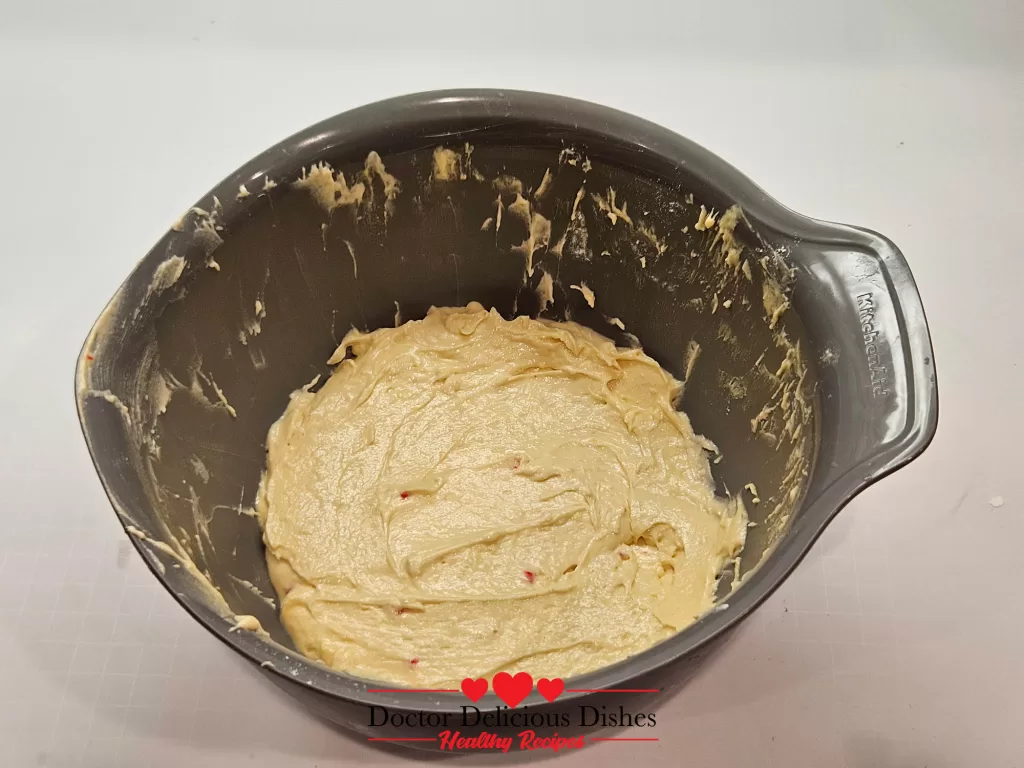 A gray mixing bowl holding smooth, creamy peppermint cake batter speckled with tiny red candy cane bits, ready for the oven.