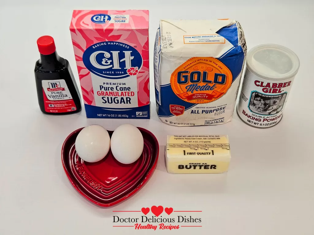 Ingredients laid out on a white surface: granulated sugar, vanilla extract, eggs in a red heart-shaped bowl, all-purpose flour, baking powder, and a stick of butter.