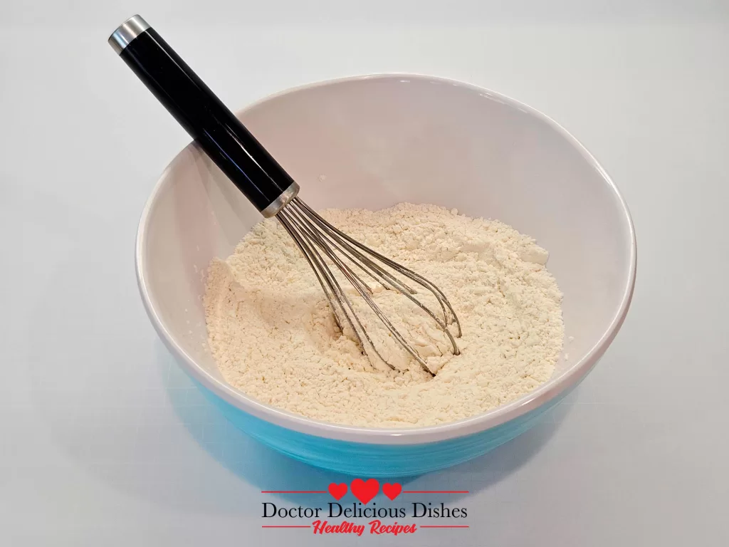 A blue mixing bowl with dry ingredients (flour and baking powder) being whisked together.