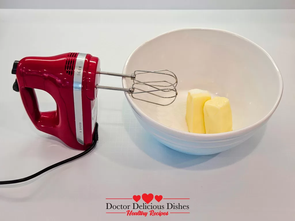 A red hand mixer beside a bowl with two sticks of butter and sugar, ready to be creamed together.