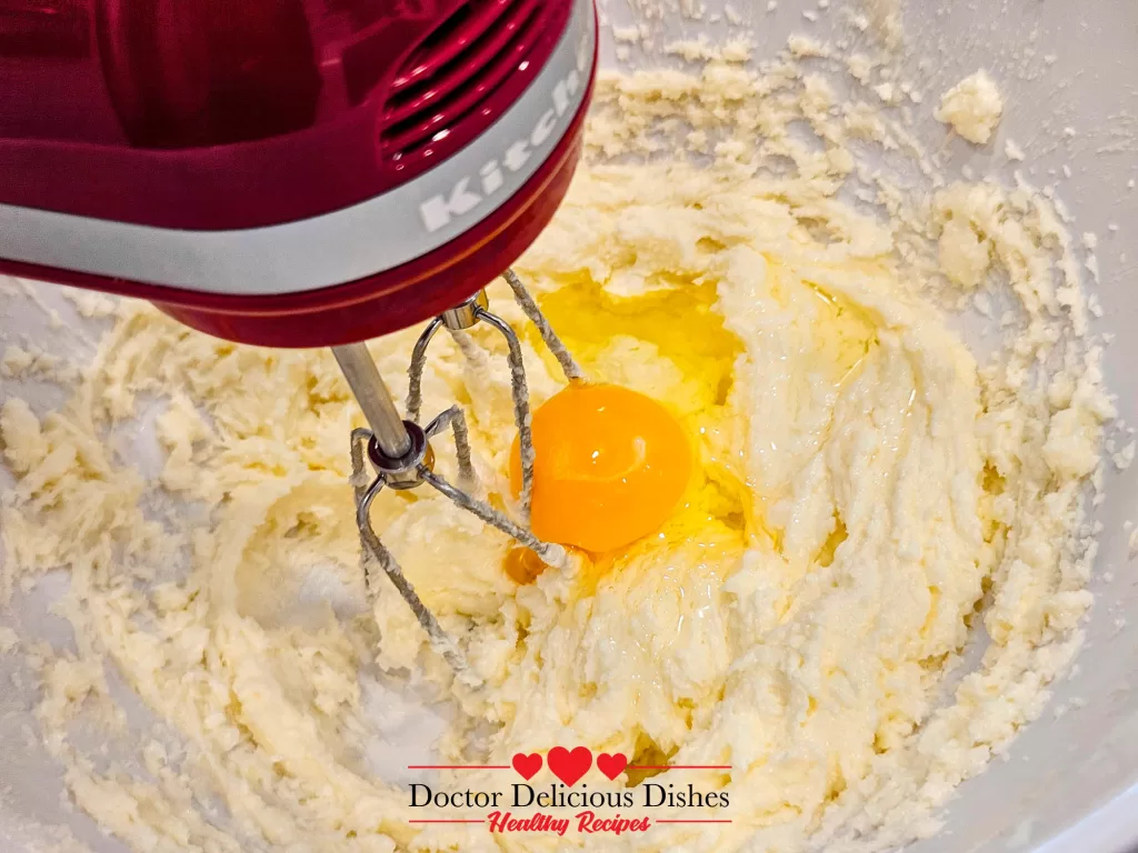 One egg being added to the creamed butter and sugar mixture, with the mixer partially visible in the process.