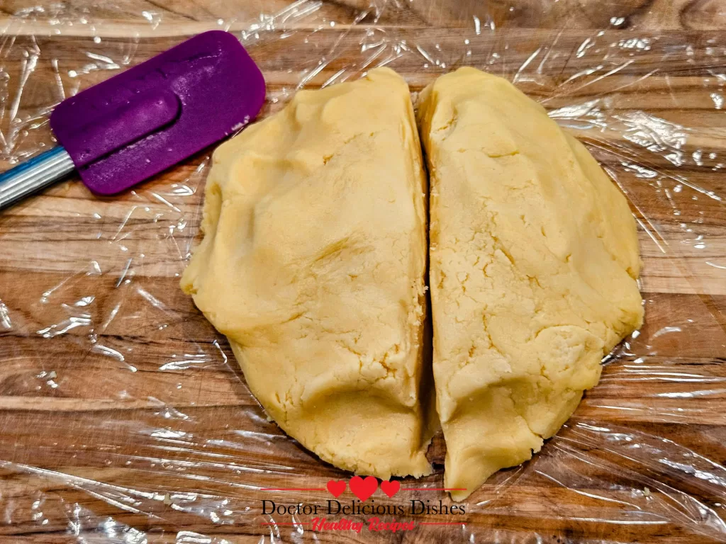 Cookie dough divided into two portions, placed on plastic wrap on a wooden surface with a purple spatula beside it.