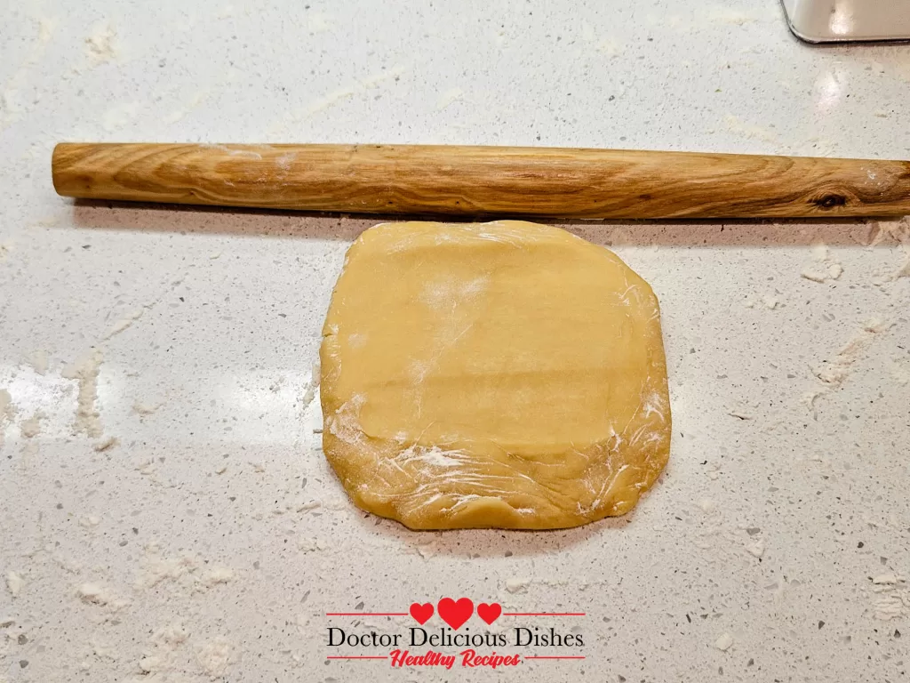 Rolled-out cookie dough on a floured countertop with a wooden rolling pin at the top.