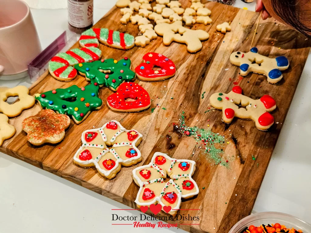 Sugar cookies being decorated on a wooden board, featuring colorful icing, sprinkles, and edible glitter. A hand is seen adding details to a cookie.