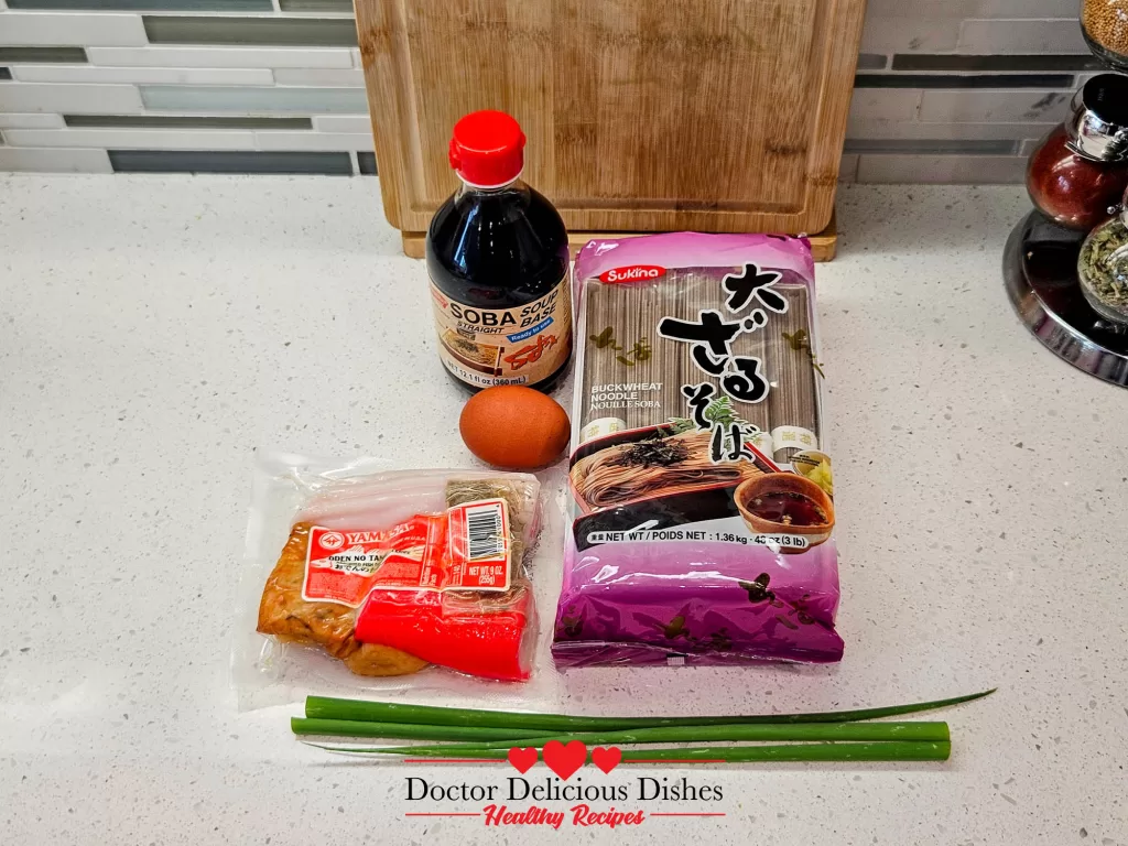 Ingredients for soba noodle soup, including soba noodles, fish cakes, soba soup base, eggs, green onions, and a cutting board on a clean kitchen countertop.