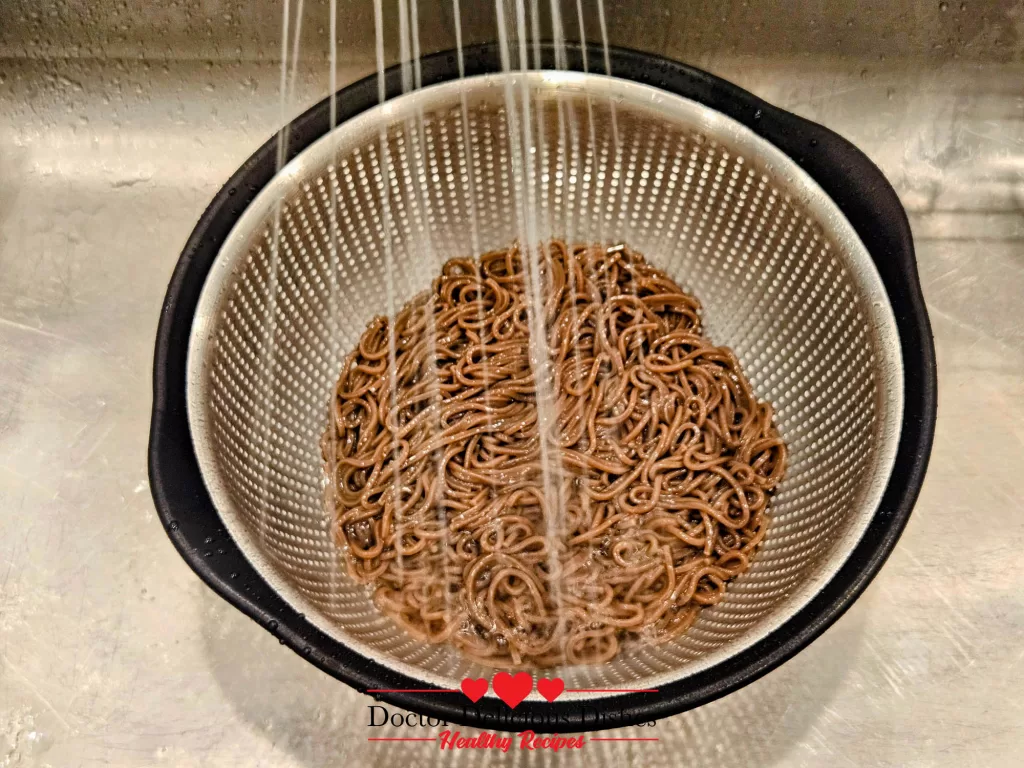 Cooked soba noodles being rinsed with cold water in a colander to remove excess starch.