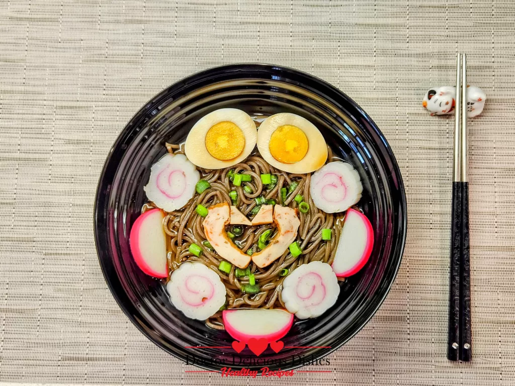 Hot soba noodle soup beautifully garnished with boiled egg halves, sliced fish cakes, and green onions in a black bowl.