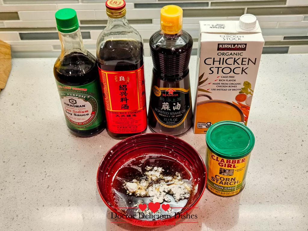 A red bowl containing sauce ingredients with cornstarch added for thickening. Bottles of soy sauce, Shaoxing wine, sesame oil, and chicken stock are visible in the background.