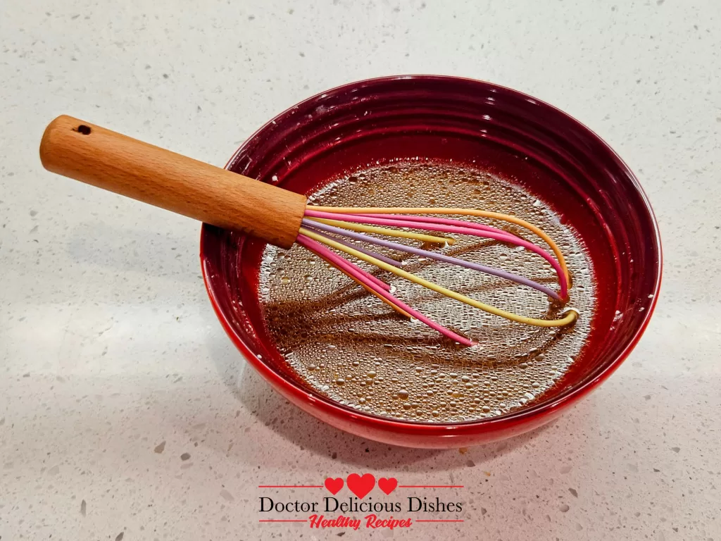 A whisk resting in a red bowl filled with a well-mixed chow mein sauce, featuring a glossy, dark brown texture ready to coat the noodles.