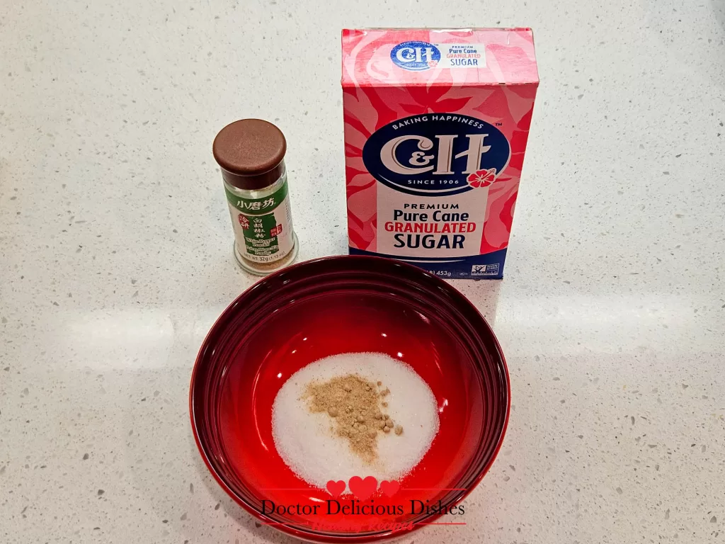 A red bowl containing a mix of white sugar and white pepper, with a box of C&H granulated sugar and a bottle of white pepper seasoning placed in the background on a countertop.