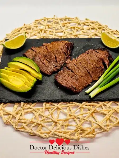 Sliced Air Fryer Skirt Steak beautifully presented with avocado, lime wedges, and green onions on a black slate plate.