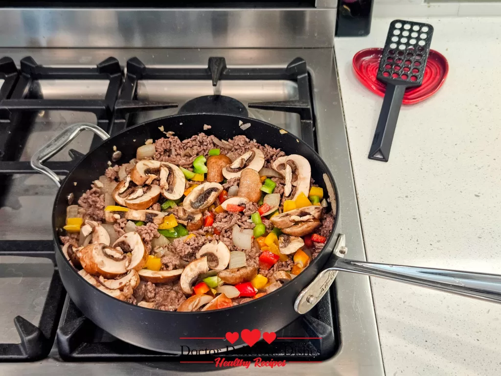 Ground beef mixture cooking in a skillet with bell peppers, onions, and mushrooms being stirred together.