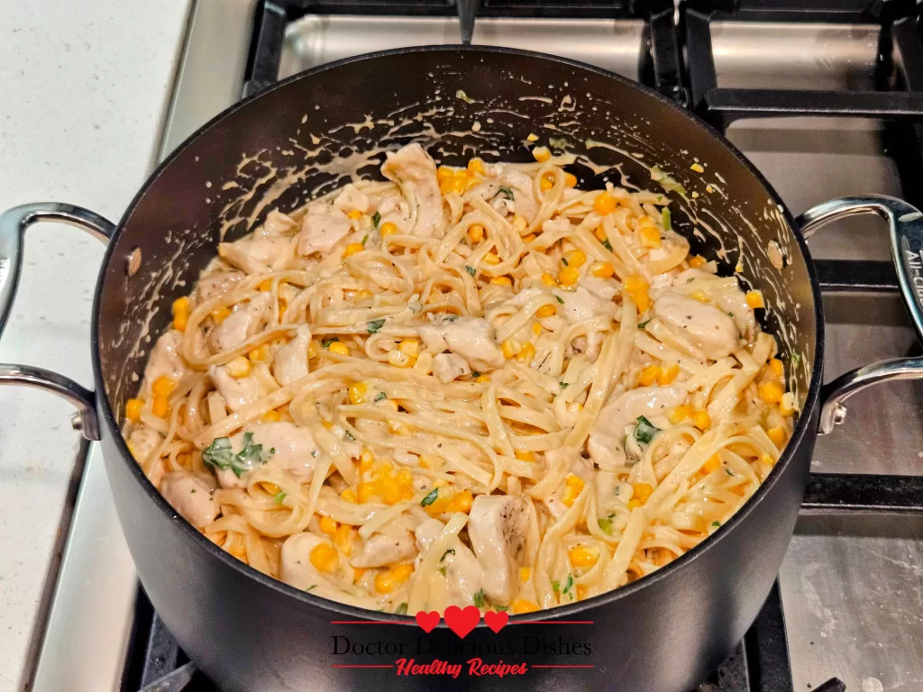 Cooked chicken, pasta, and corn being stirred into the Alfredo sauce in a large pot for a Chicken Alfredo Recipe with Jar Sauce.