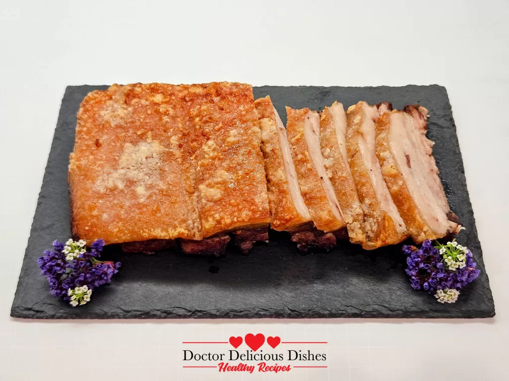 Golden, crispy pork belly sliced and served on a black slate platter, garnished with edible flowers.