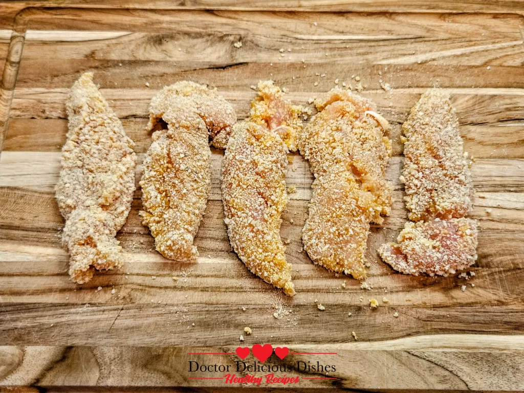 Freshly breaded Crispy Air Fryer Chicken Tenders lined up on a wooden cutting board, ready for air frying.